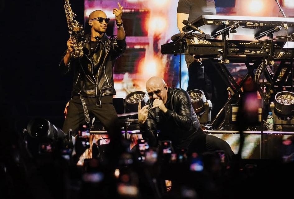 Luiso points to the crowd with his saxophone in hand while Pitbull sings at the front of the stage.
