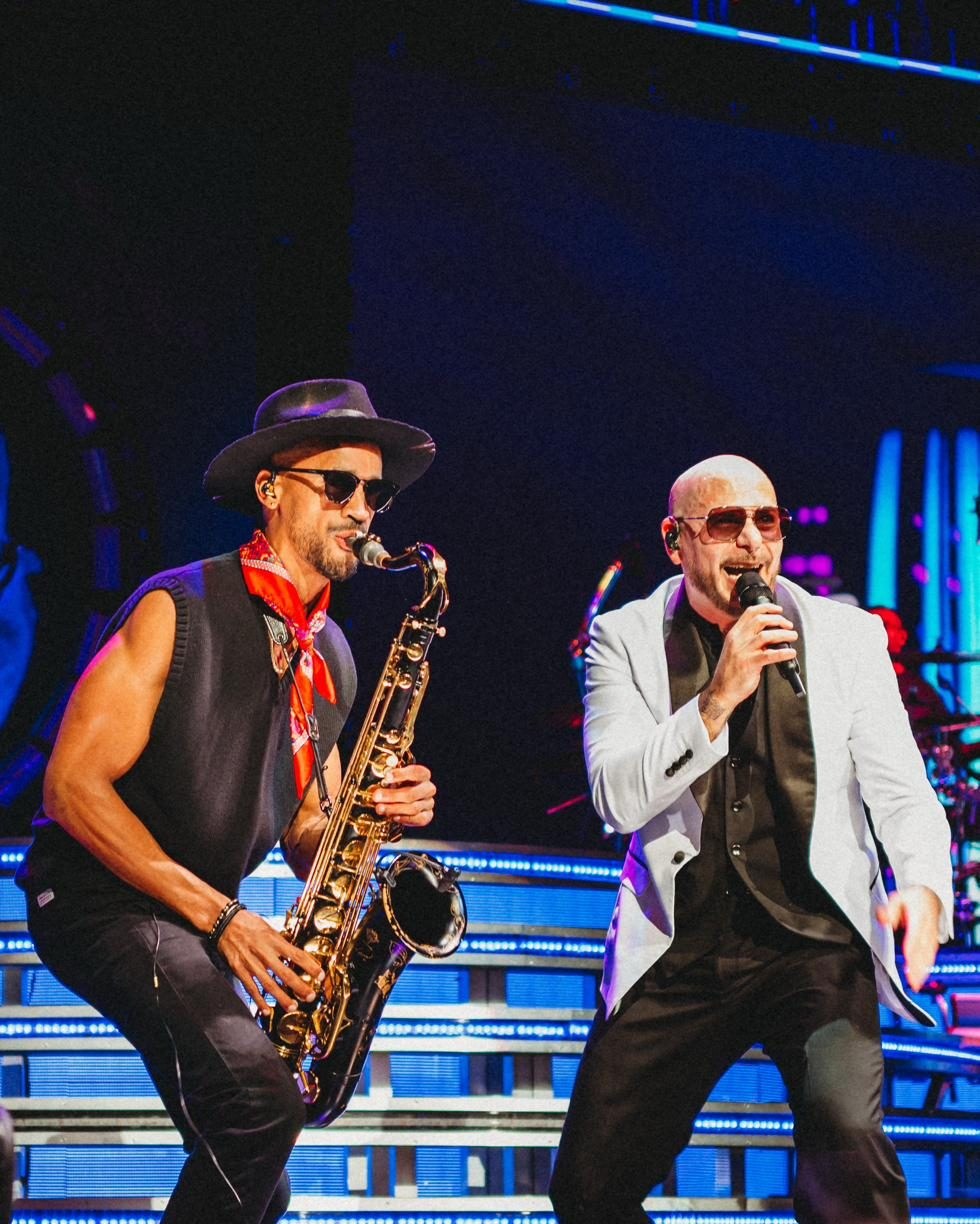 Luiso on saxophone beside Pitbull under blue lights, wearing a hat and a red bandana.