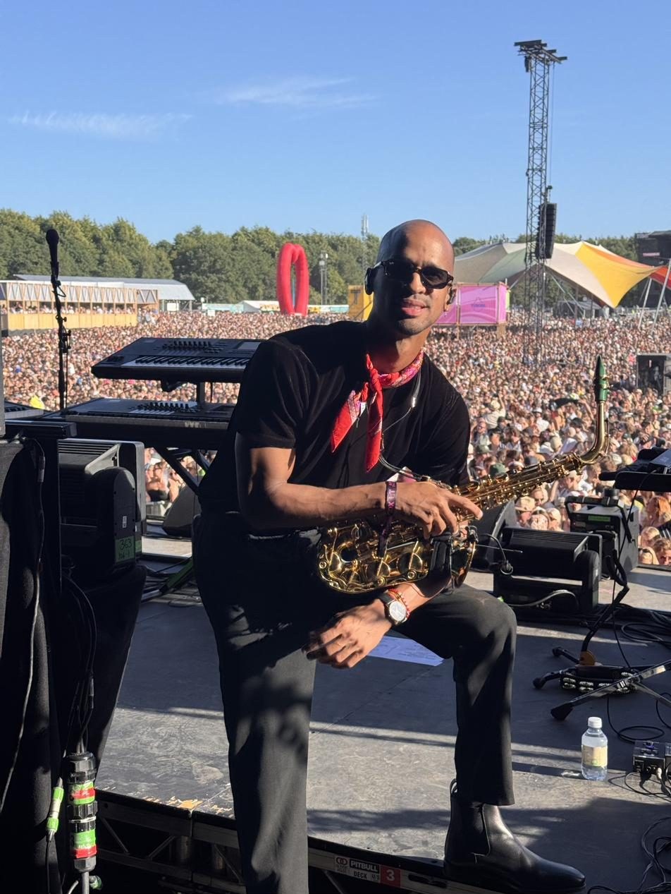 Luiso sitting on the edge of the stage with his saxophone, the festival crowd behind him.