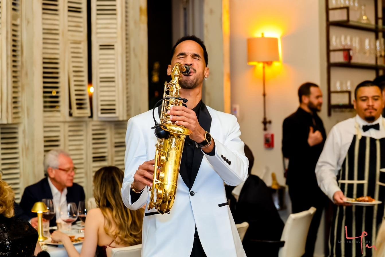 Luiso playing saxophone in a white tuxedo among the tables of a restaurant.