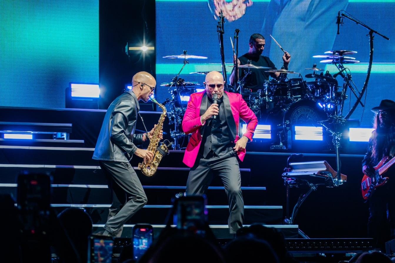 Luiso playing saxophone next to Pitbull in concert, with the drummer behind them.
