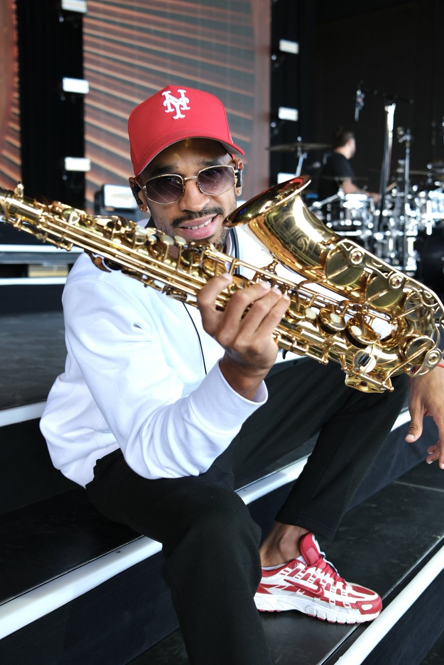 Luiso sitting on the stage steps in a red cap and sunglasses, holding his alto saxophone and smiling at the camera.